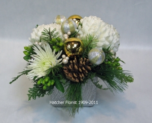 A silver ceramic shimmering ceramic base. White Commercial mums and Anastasia mums, two tone gold balls, cones, seasonal pine and fir. let our elves deliver one for you.