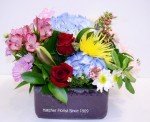 A purple ceramic square container holds a garden of fresh Sierra Eco flowers. A mauve butterfly is nestled amongst the hydrangea, roses, anastasia mums, alstromeria, assorted greenery.